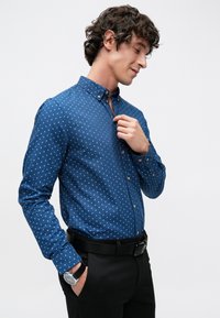 Sinine nööpidega särk, valge ruudumustriga, pikkade varrukatega ja kraega. Kantud mustade pükstega ja hõbedase käekellaga.