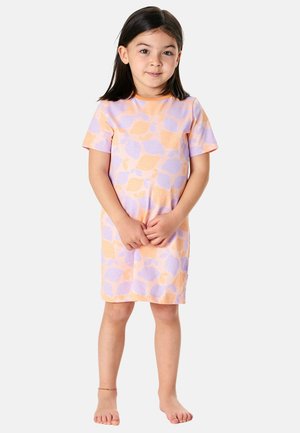 Young girl with dark hair wearing a pastel-colored lemon-patterned dress, standing barefoot against a plain light background.