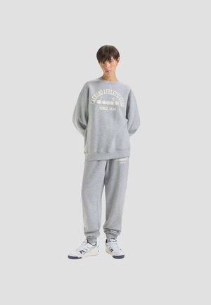 Completo felpa grigia con pantaloni della tuta, caratterizzato da un collo rotondo, vestibilità ampia e stampa grafica frontale. Materiale morbido, con una texture simile al pile.