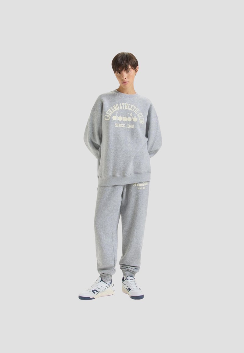 Completo felpa grigia con pantaloni della tuta, caratterizzato da un collo rotondo, vestibilità ampia e stampa grafica frontale. Materiale morbido, con una texture simile al pile.