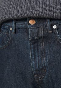 Jeans de denim azul oscuro con un botón de cobre, costuras visibles y una etiqueta que dice "CLOSED". Un suéter de punto gris cubre parcialmente la cintura.