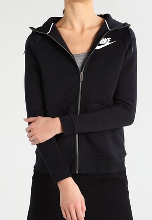 Osoba nosząca czarną bluzę z kapturem na zamek Nike na szarej koszulce, z rękami skrzyżowanymi na wysokości talii na jednolitym tle.