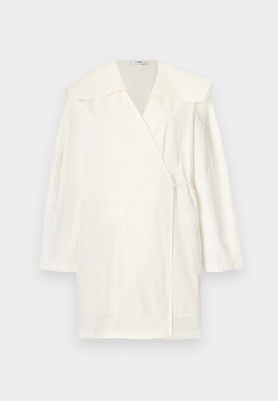 Λευκό παλτό με μακριά μανίκια και τύλιγμα, με φαρδιά γιακά, δύο μπροστινές τσέπες και μια μίνιμαλ δέσιμο στο πλάι, προβαλλόμενο σε απλό φόντο.