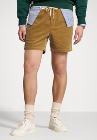 Man som bär bruna kordflorsshorts med dragsko, beige strumpor och vita sneakers, med en randig skjortslut och gröna sweatshirtärmar synliga.