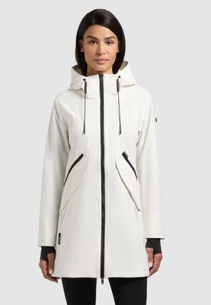 Vrouw draagt een witte jas met capuchon, zwarte rits en zakdetails, staand tegen een effen lichtgrijze achtergrond.