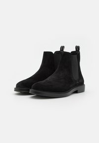Bottines Chelsea en suède noir avec des panneaux élastiques sur les côtés et une semelle en caoutchouc plate. Comprend des languettes de tirage pour un enfilage facile, une texture lisse et un design classique.