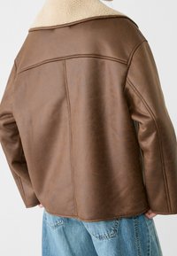 Veste en simili cuir marron avec col en shearling blanc portée sur un jean en denim bleu clair, vue de dos.