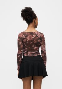 Γυναίκα με σγουρά μαλλιά, ντυμένη με μπλούζα μακρυμάνικη με λουλουδάτο μοτίβο και μαύρη φούστα με πλισέ, στέκεται με την πλάτη προς τα εμπρός σε απλό λευκό φόντο.