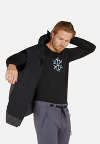 Svart långärmad skjorta för män med ett blått mönster av snöflingor, i kombination med en mörk kviltad väst och ljusgrå byxor som har dragkedjefickor.