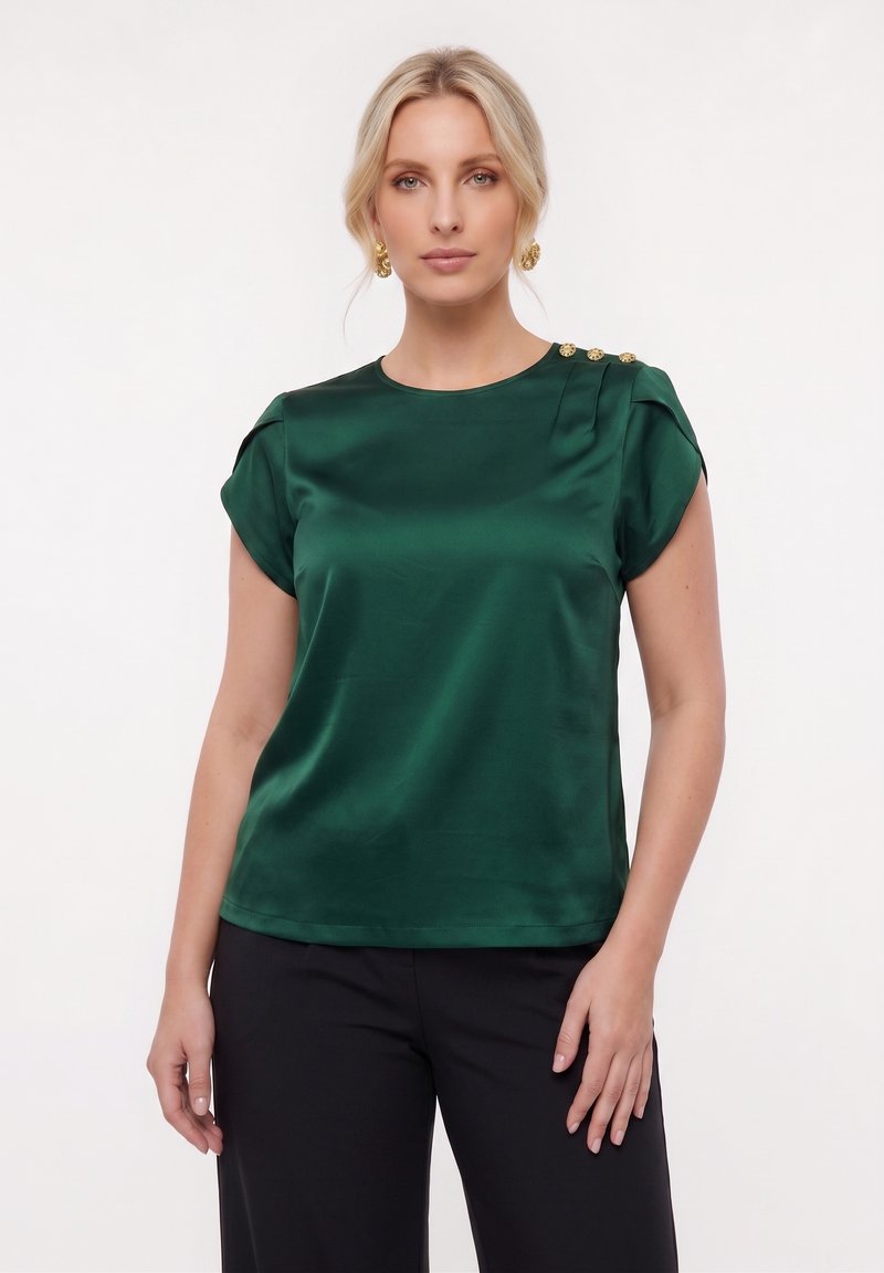 Vrouw draagt een donkergroene satijnen top met korte mouwen met gouden knopen op de schouder en zwarte broek tegen een witte achtergrond.