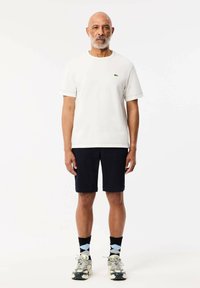 Maglietta in cotone bianco a maniche corte con un sottile logo verde; abbinata a pantaloni blu navy. Scarpe da ginnastica grigie e nere, calze a fantasia.