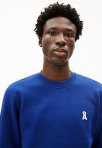 Blauwe sweatshirt met een ronde hals, geribbelde kraag en een wit geborduurd logo op de linkerborst. Zachte textuur, casual ontwerp.