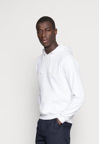 Witte hooded sweatshirt met een voorzak en trekkoord. Stof lijkt zacht; model draagt donkere broek en heeft kort haar.