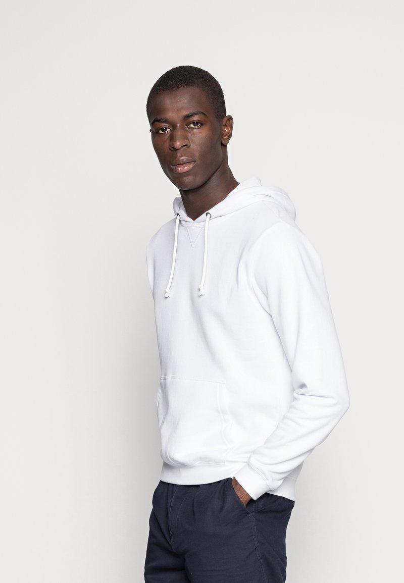 Witte hooded sweatshirt met een voorzak en trekkoord. Stof lijkt zacht; model draagt donkere broek en heeft kort haar.