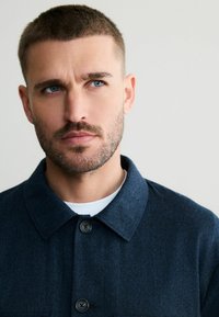 Giacca button-up blu navy realizzata in tessuto testurizzato, con colletto e due bottoni scuri sul davanti.