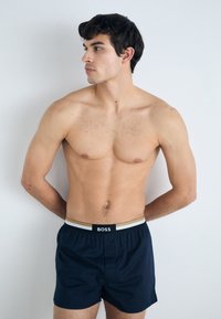 Boxers bleu marine avec une ceinture élastique noire et dorée arborant le logo "BOSS". Le tissu semble léger et lisse.