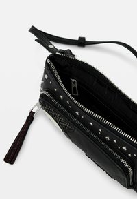 Bolso de cuero negro con tachuelas plateadas y cierre de cremallera. Paneles texturizados con un patrón tejido. Correa desmontable con costuras rojas.