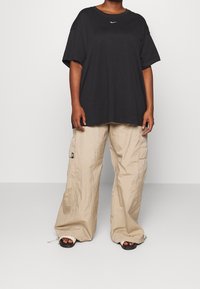 Person som bär en överdimensionerad svart Nike-t-shirt,vida beige cargo-byxor och svarta slip-on sandaler med krämfärgade remmar, stående på en grått golv.