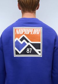 Felpa blu con un grande logo rettangolare sul retro. Il logo include grafiche di montagne in arancione, nero e blu con il testo "NAPAPIJRI".
