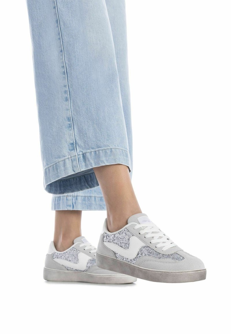 Zapatillas grises y blancas con acentos brillantes, acabado de ante, paneles laterales texturizados y suela de goma, combinadas con vaqueros de pierna ancha de color azul claro.