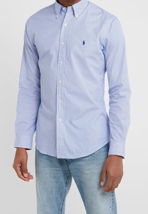 Mann trägt ein hellblau-weiß gestreiftes, dünnes Hemd mit langen Ärmeln und hellblaue Jeans, steht vor einem einfarbigen Hintergrund.