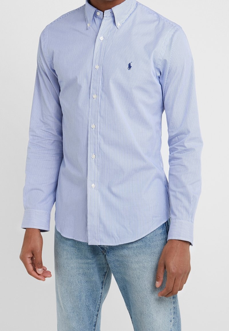Homme portant une chemise boutonnée à fines rayures bleu clair et blanc à manches longues et un jean bleu clair, debout devant un fond uni.