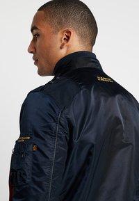 Navy bomber jacket in shiny nylon material, featuring ribbed collar, zipper closure, and side pocket with orange accent.
