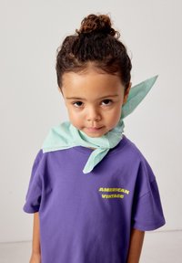 Enfant aux cheveux bouclés attachés en chignon, portant un t-shirt violet "American Vintage" et une écharpe vert clair nouée autour du cou, face à l'avant.