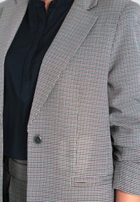 Marynarka w kratę w jasnoszarym, czerwonym i czarnym odcieniach. Posiada klapę, zapięcie na jeden guzik oraz dwie przednie kieszenie.