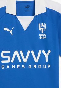 Mėlynas trumpomis rankovėmis sportinis marškinėlis su baltu apykakle. Puošia baltas Puma logotipas, „ALHILAL“ logotipas ir „SAVVY GAMES GROUP“ drąsiu baltu tekstu.