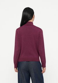 Mulher com cabelo preto comprido usa um suéter roxo de gola alta ajustado e calças largas azul-escuro, de costas contra um fundo branco.