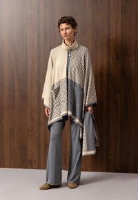 Femme debout contre un mur en panneaux de bois, portant une cape beige et grise à motif pied-de-poule avec un pantalon gris et des chaussures marron.