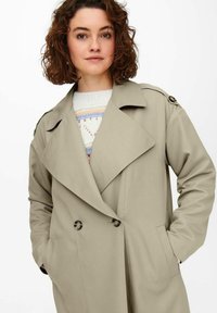 Beige Trenchcoat mit doppelter Knopfleiste, großen Revers und Seitentaschen. Verfügt über dekorative Knöpfe an den Schultern und eine glatte Stoffoberfläche.