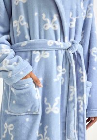 Jasnoniebieski szlafrok z polaru z białymi wzorami kokardek, przewiązywany w talii, z dwoma przednimi kieszeniami, wykonany z miękkiego, teksturowanego materiału.