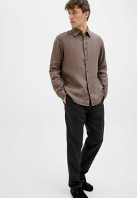 Brown linen long-sleeve shirt with button closure, worn with black pants and dark shoes. Plain design with a relaxed fit and collar.