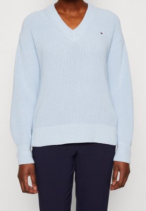 Personne portant un pull en laine bleu clair à col en V avec manches longues et pantalon foncé, visible des épaules aux hanches.