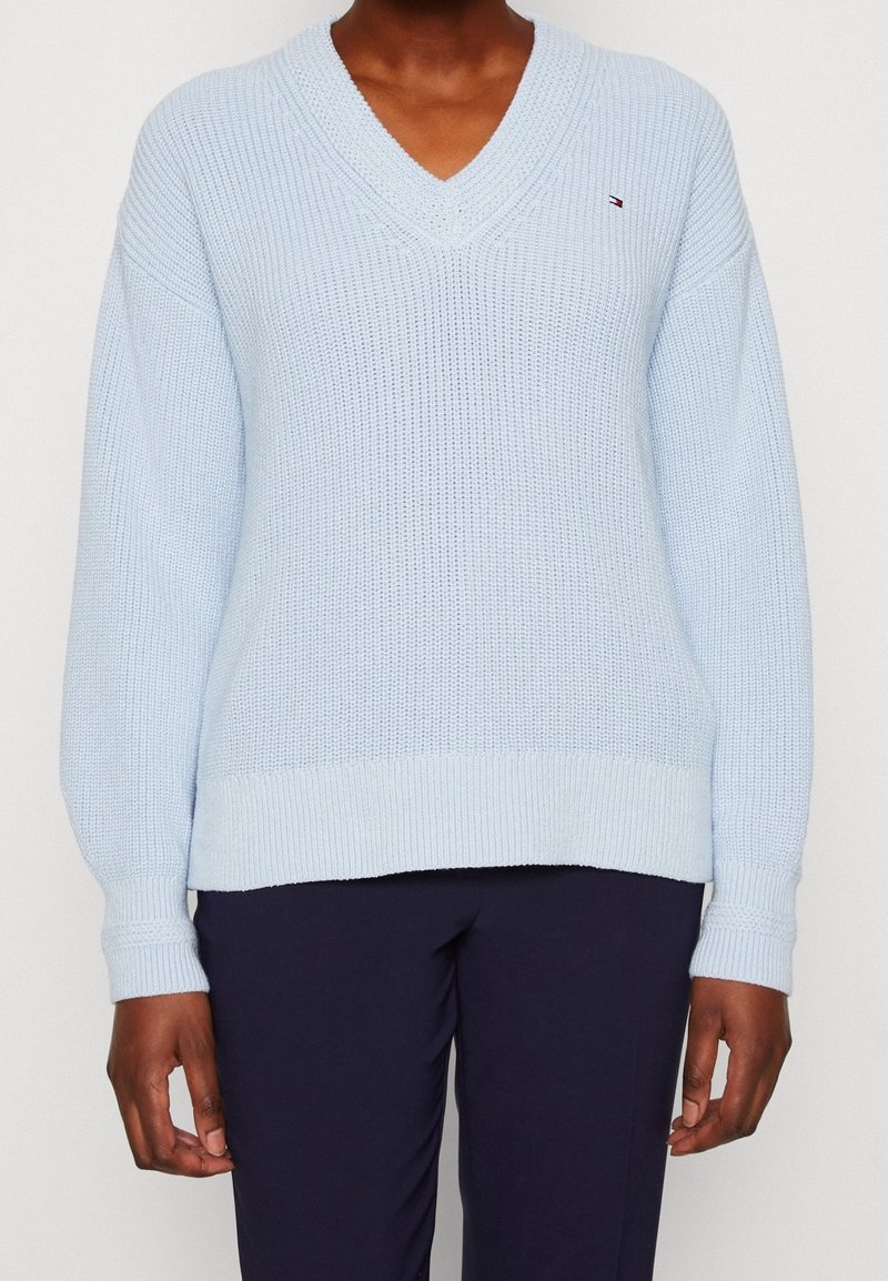 Personne portant un pull en laine bleu clair à col en V avec manches longues et pantalon foncé, visible des épaules aux hanches.