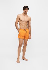 Jeune homme debout, pieds nus, portant un short de bain orange avec un cordon noir sur un fond blanc uni.