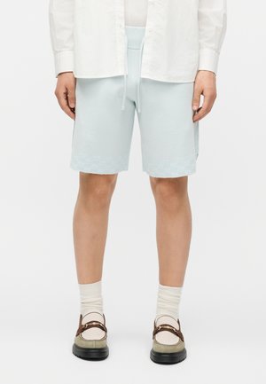 Άνδρας που φοράει ανοιχτό μπλε σορτς με κορδόνι, λευκό πουκάμισο, κρεμ κάλτσες και μπεζ loafers με καφέ λουριά σε απλό λευκό φόντο.