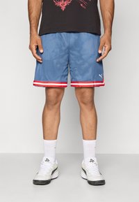 Blå mesh idrettsshorts med rød og hvit stripet kant, sidelommer og hvit Puma-logo. Brukt med hvite sokker og sneakers.