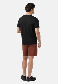Mand står med ryggen til, iført sort t-shirt, rustfarvede cargo-shorts og sorte sneakers foran en ensfarvet baggrund.
