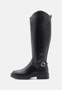 Černá kožená vysoká bota a�ž ke kolenům s embosovaným geometrickým vzorem na zadní straně, zlatou kulatou přezkou blízko kotníku a nízkým blokovým podpatkem.