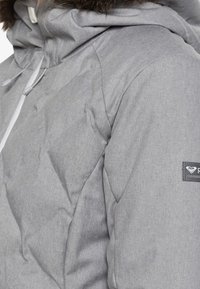 Graue Pufferjacke mit Kapuze und Fellbesatz, strukturiertem Stoff und einem Logo-Patch am Ärmelsaum. Sichtbare Reißverschluss- und Kordelzugdetails.