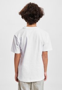 Barn med lockigt mörkt hår som bär en enkel vit t-shirt och ljusa byxor, stående vänd bort från kameran mot en vit bakgrund.