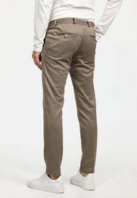 Pantalons marron ajustés en tissu texturé, présentant une coupe slim, deux poches arrière avec boutons et un motif subtil. Associés à des baskets blanches.