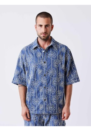 Homme aux cheveux courts et barbe, portant une chemise et un pantalon bleus assortis avec des motifs floraux délimités en blanc, debout devant un fond uni.