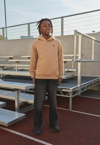 Tanfarvet hættetrøje med en lille kranielogo, kombineret med løse grå jeans og sorte sko. Baggrunden viser metalliske tilskuerrækker og en løbebane.