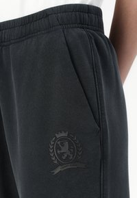 Tamsiai pilki sportiniai kelnes su elastinga juosta, šoninėmis kišenėmis ir toniniu logotipu, kuriame vaizduojamas liūtas su laurų vainiku ant šlaunies.