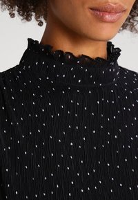 Black textured fabric top with a ruffled collar, featuring scattered white dots, creating a pleated effect. Close-up detail of neckline.