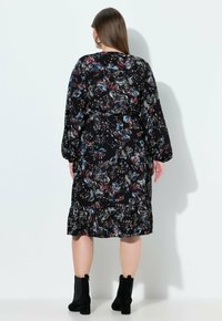 Musta mekko, jossa on värikäs perhonen ja kukkakuvio, pitkät hihat, joustava vyötärö ja kerroksittainen helma, yhdistetty mustiin nilkkureihin.